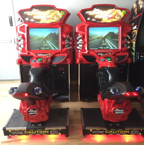 Two red motorcycle arcade games with screens displaying a racing scene, set on a wooden floor.