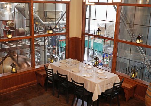A dining table set for a meal, with large windows showing a brewery in the background. Warm lighting and wooden decor.