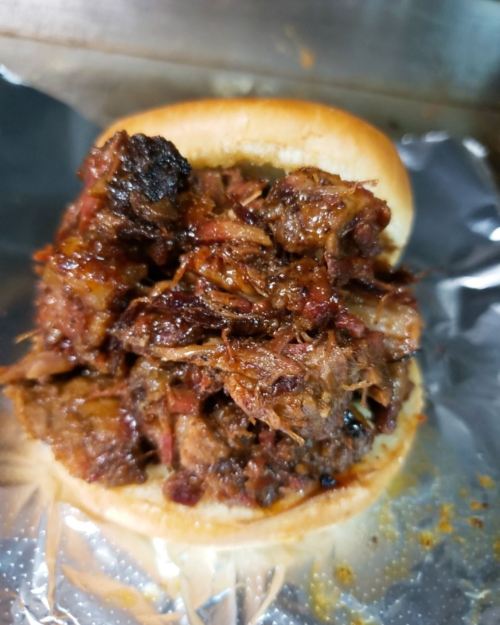 A close-up of a sandwich filled with shredded, smoky barbecue meat on a soft bun, resting on foil.
