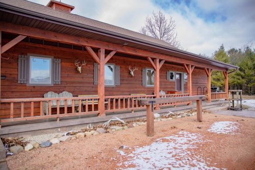 A rustic wooden cabin with a porch, featuring antler decor and a gravel pathway, surrounded by trees and snow.