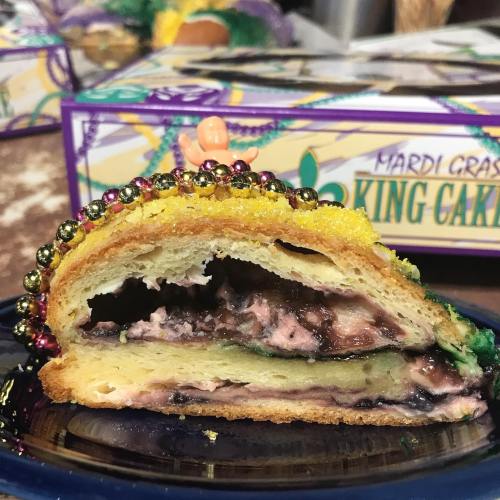 A slice of King Cake with colorful icing, beads, and a creamy filling, set against a festive background.