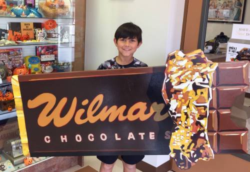 A young boy smiles while holding a large, colorful sign for Wilmar Chocolates in a candy shop.