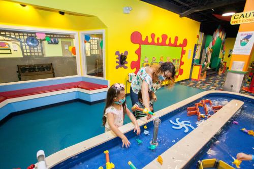 A child and an adult play with water toys at a colorful indoor play area. Bright walls and fun decorations are visible.