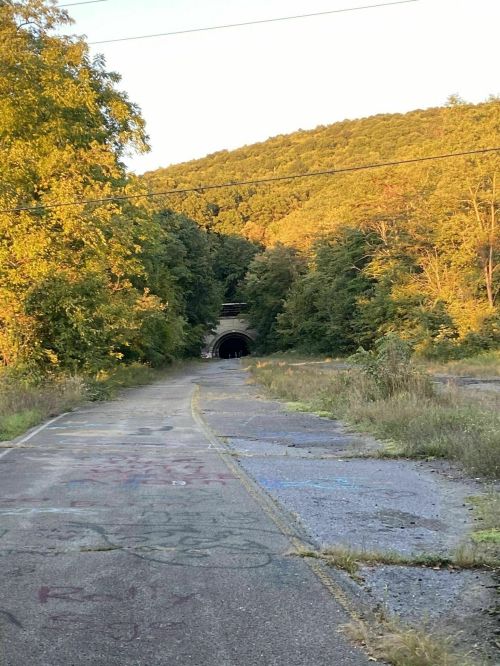A deserted road leads to a dark tunnel, surrounded by trees and hills, with graffiti on the pavement.