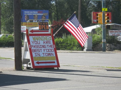 A roadside sign reads "WARNING: YOU ARE PASSING THE BEST FOOD IN TOWN," with an American flag in the background.