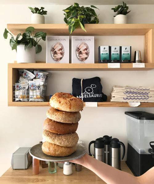 A hand holds a plate stacked with various bagels in front of a shelf with plants and products.