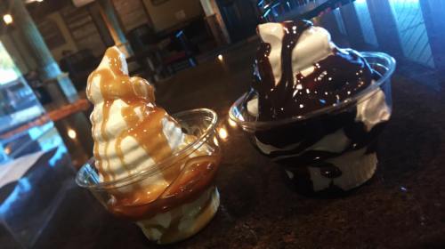 Two cups of soft serve ice cream: one topped with caramel sauce and the other with chocolate sauce, on a dark countertop.