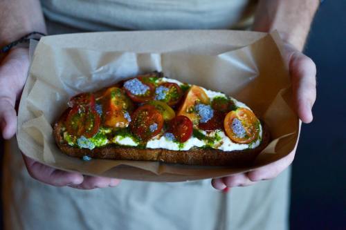 A person holds a slice of toast topped with tomatoes, cream, and green pesto, served in a paper tray.