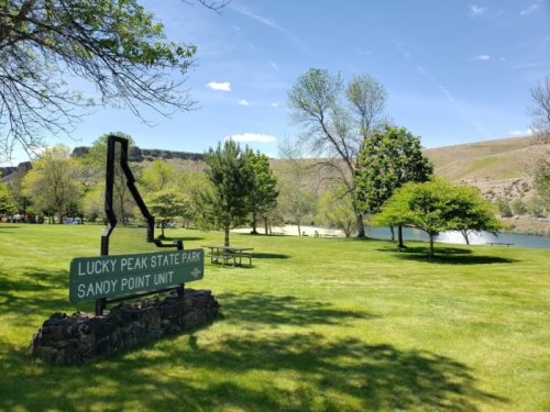 Sign for Lucky Peak State Park's Sandy Point Unit, with green grass, trees, and a lake in the background.