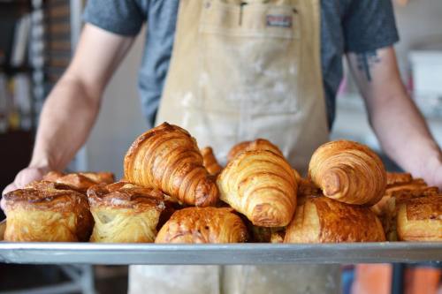 A person in an apron holds a tray filled with various freshly baked pastries, including croissants and danishes.
