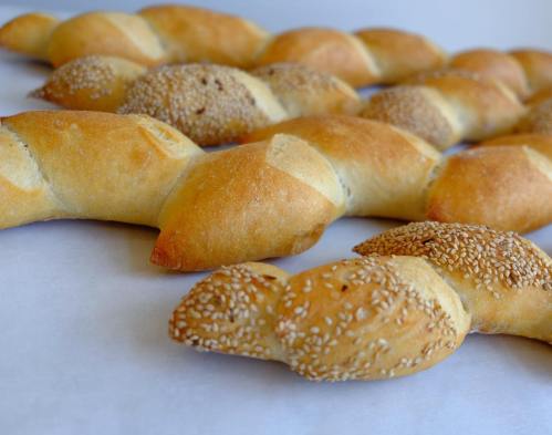 A close-up of freshly baked twisted bread rolls, some topped with sesame seeds, arranged on a white surface.