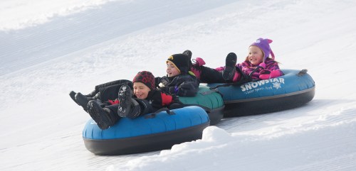 Three children joyfully tubing down a snowy slope, wearing colorful winter gear and laughing together.