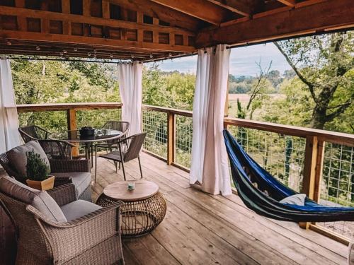 Cozy outdoor deck with a hammock, table, chairs, and curtains, surrounded by lush greenery and a scenic view.