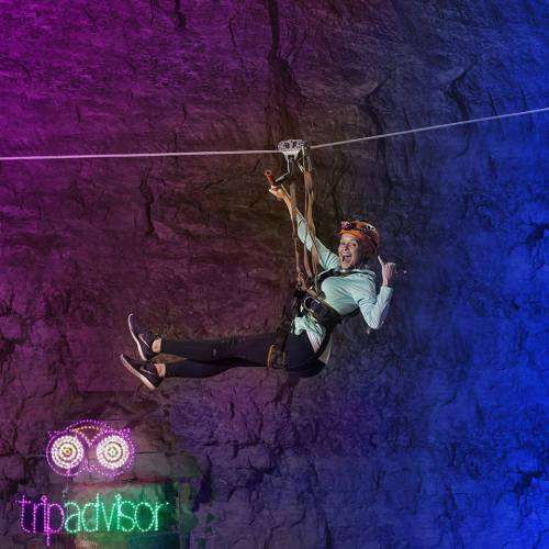 A person zip-lining in a cave, wearing a helmet and harness, with colorful lighting and a TripAdvisor logo below.
