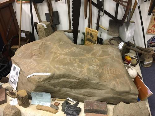 A large rock with ancient carvings, surrounded by various historical tools and artifacts in a museum setting.
