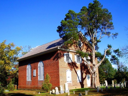 Historic brick church surrounded by a cemetery and tall trees under a clear blue sky.
