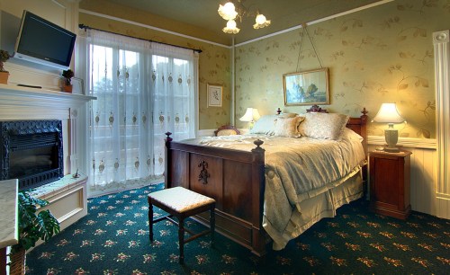 Cozy bedroom with a wooden bed, fireplace, and soft lighting, featuring floral wallpaper and a view through sheer curtains.