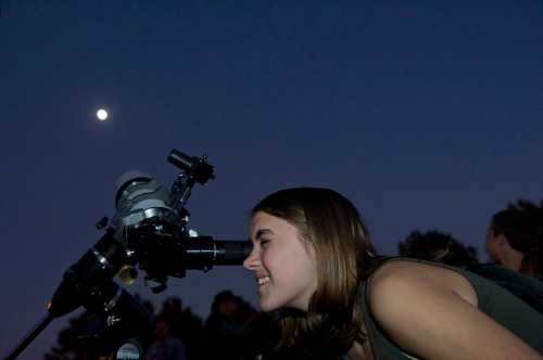 A young woman smiles while looking through a telescope at the night sky, with the moon visible above.