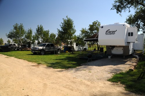 A gravel road leads to a campsite with an RV, parked trucks, and trees under a clear blue sky.
