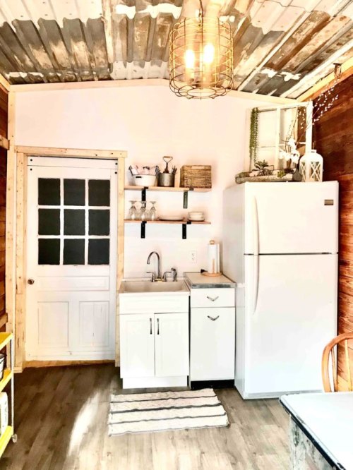 A cozy kitchen featuring a white fridge, sink, shelves, and rustic decor with wooden walls and a metal ceiling.