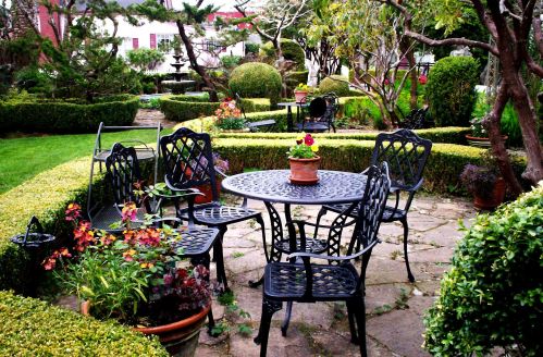A serene garden scene featuring a black metal table and chairs surrounded by lush greenery and colorful flowers.