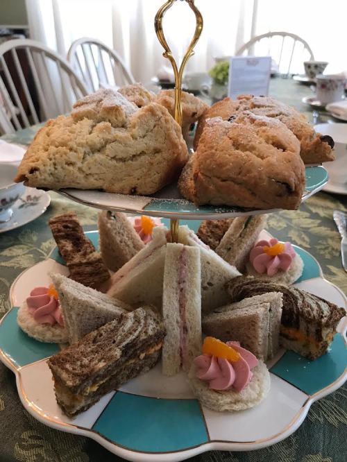 A tiered stand with scones on top and assorted finger sandwiches decorated with pink flowers on the bottom.
