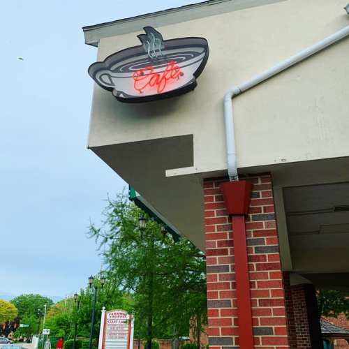 A café sign with a steaming cup logo, mounted on a brick building under a cloudy sky.