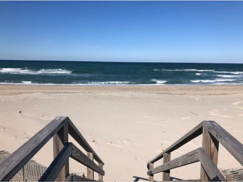 View of a sandy beach with wooden stairs leading down to the ocean, under a clear blue sky. Waves gently lap at the shore.