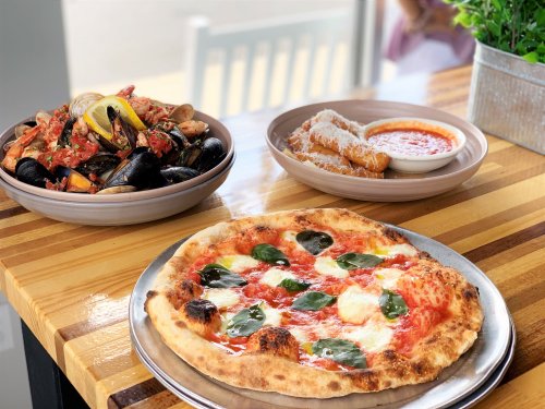 A table with a pizza topped with basil, a seafood dish, and fried food with dipping sauce, all on rustic wooden surfaces.