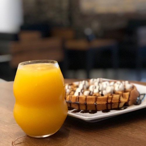 A glass of orange juice next to a plate of waffles topped with chocolate and marshmallows on a wooden table.