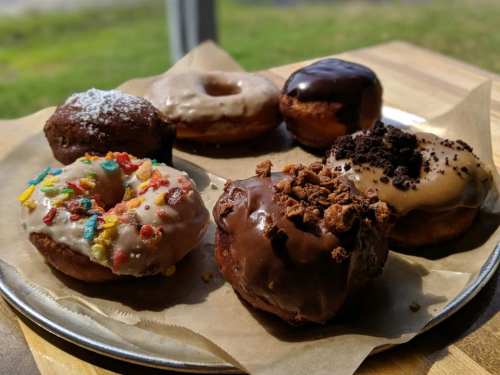 A plate of assorted donuts, including chocolate, glazed, and colorful cereal-topped varieties, on a wooden table.