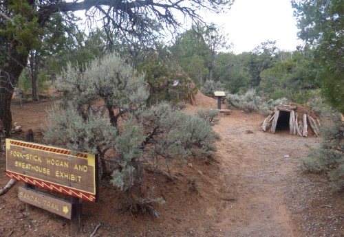 Sign for "Fork-Stick Hogan and Sweathouse Exhibit" near a small structure, surrounded by shrubs and trees on a dirt path.