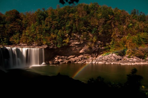 A serene waterfall surrounded by colorful autumn foliage, with a faint rainbow arching over the calm water below.