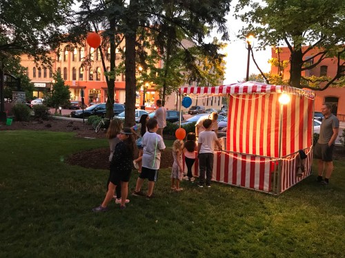 Children gather around a striped carnival booth in a park during sunset, with balloons and trees in the background.
