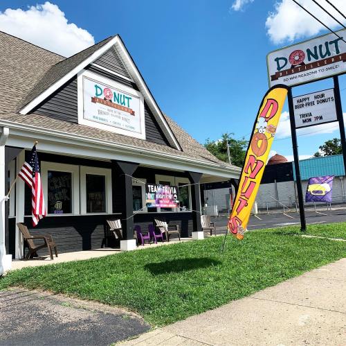 A donut shop with a colorful sign, outdoor seating, and a flag advertising donuts, set against a blue sky.
