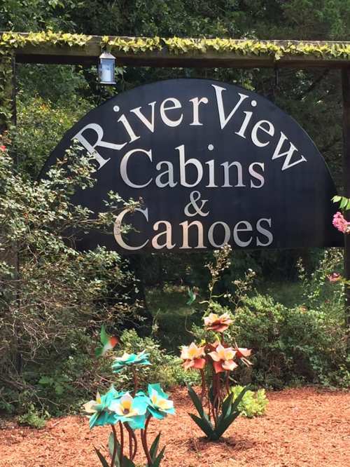 Sign for "RiverView Cabins & Canoes" surrounded by colorful flowers and greenery.