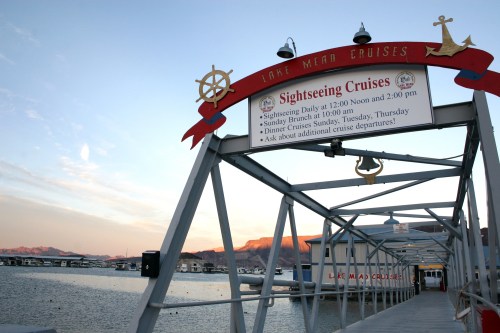 A dock at Lake Mead with a sign for sightseeing cruises and a scenic view of the water and mountains at sunset.