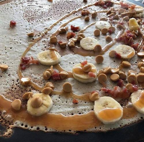 A close-up of a pancake topped with banana slices, chocolate chips, and drizzled with syrup.