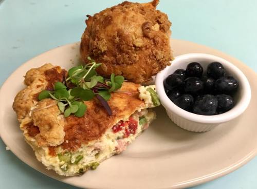 A plate with a slice of savory quiche, a muffin, and a small bowl of fresh blueberries.