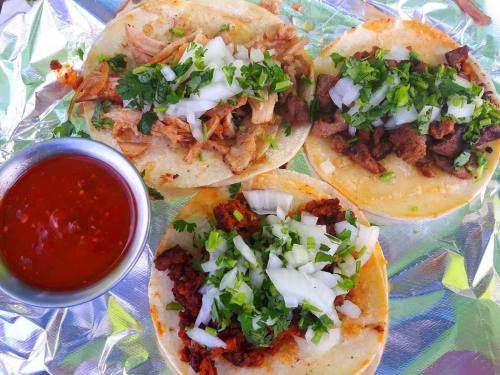 Three tacos on a foil-lined plate, topped with onions and cilantro, alongside a small bowl of red salsa.