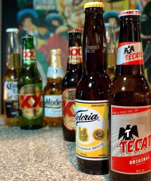 A variety of Mexican beer bottles lined up on a countertop, including Victoria, Tecate, and Modelo.