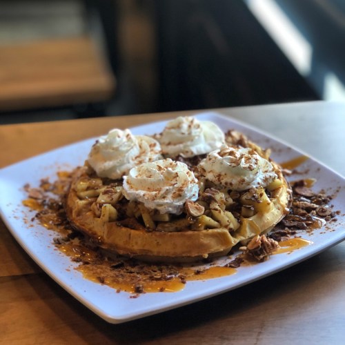 A delicious waffle topped with spiced apples, whipped cream, and drizzled with caramel on a white plate.