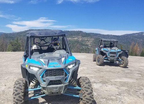 Two blue off-road vehicles parked on a rocky terrain with a mountainous landscape and clear blue sky in the background.