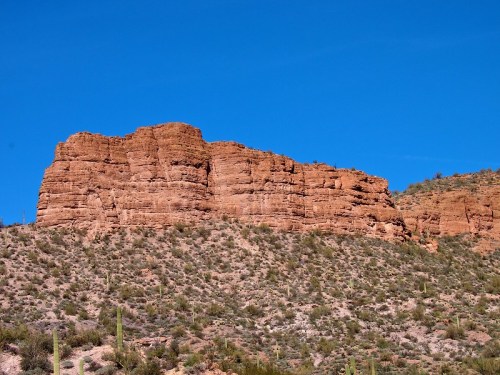 A rocky desert landscape with a prominent red rock formation under a clear blue sky. Cacti dot the foreground.