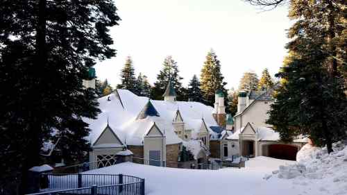 A snow-covered mansion surrounded by tall evergreen trees, with a clear sky in the background.