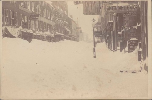Historic street scene buried in deep snow, with storefronts and signs partially visible in a winter storm.