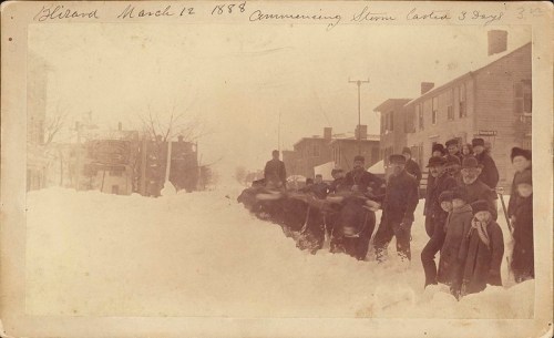 Historic photo from March 12, 1888, showing a snowy street with people and horse-drawn sleds in a winter scene.
