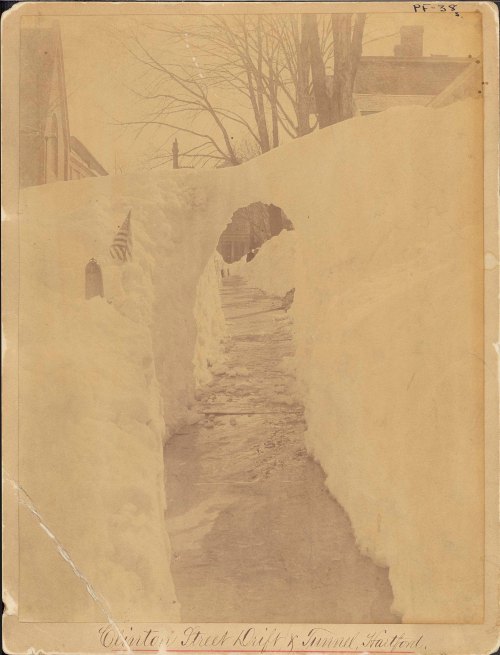 A snow-covered pathway with tall snowbanks on either side, leading through an archway of snow. Historical photo.