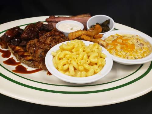 A plate featuring barbecue meats, macaroni and cheese, corn, fried onion rings, and sides of sauce and greens.
