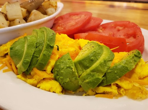 A plate featuring scrambled eggs with cheese, sliced avocado, fresh tomato, and a side of diced potatoes.